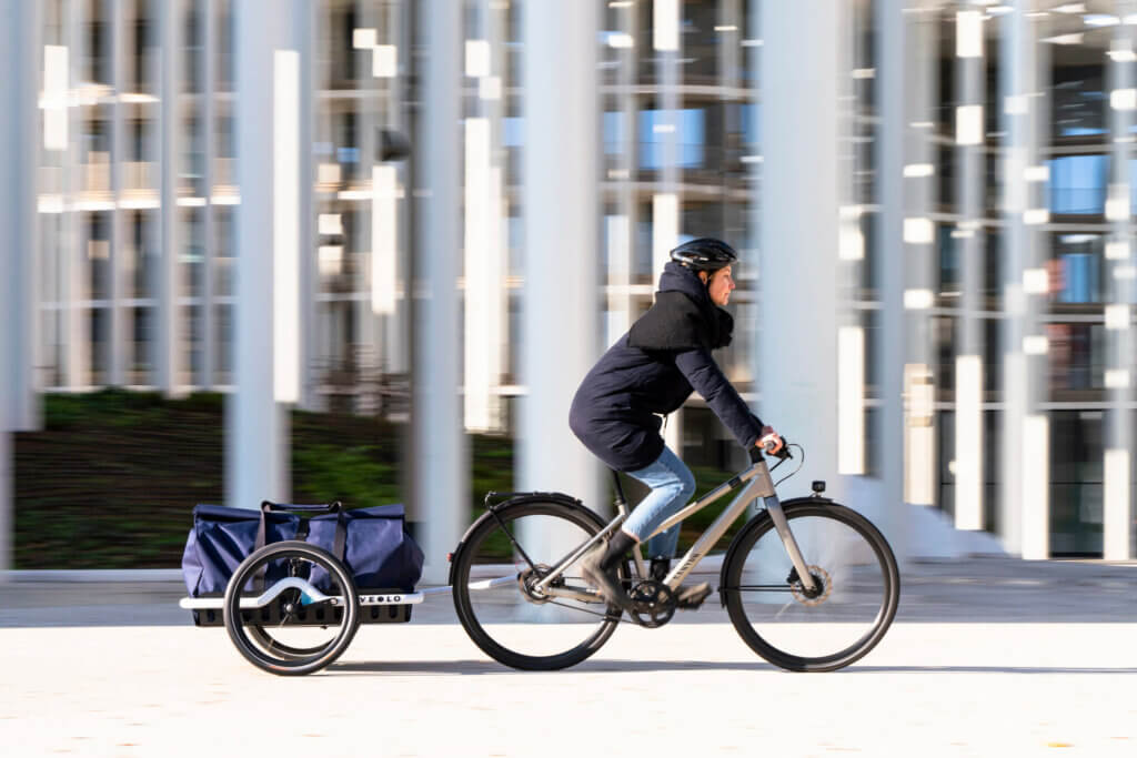 Schwarz gekleidete Fahrradfahrerin mit blauem Anhänger vor einer hellen Häuserfront.